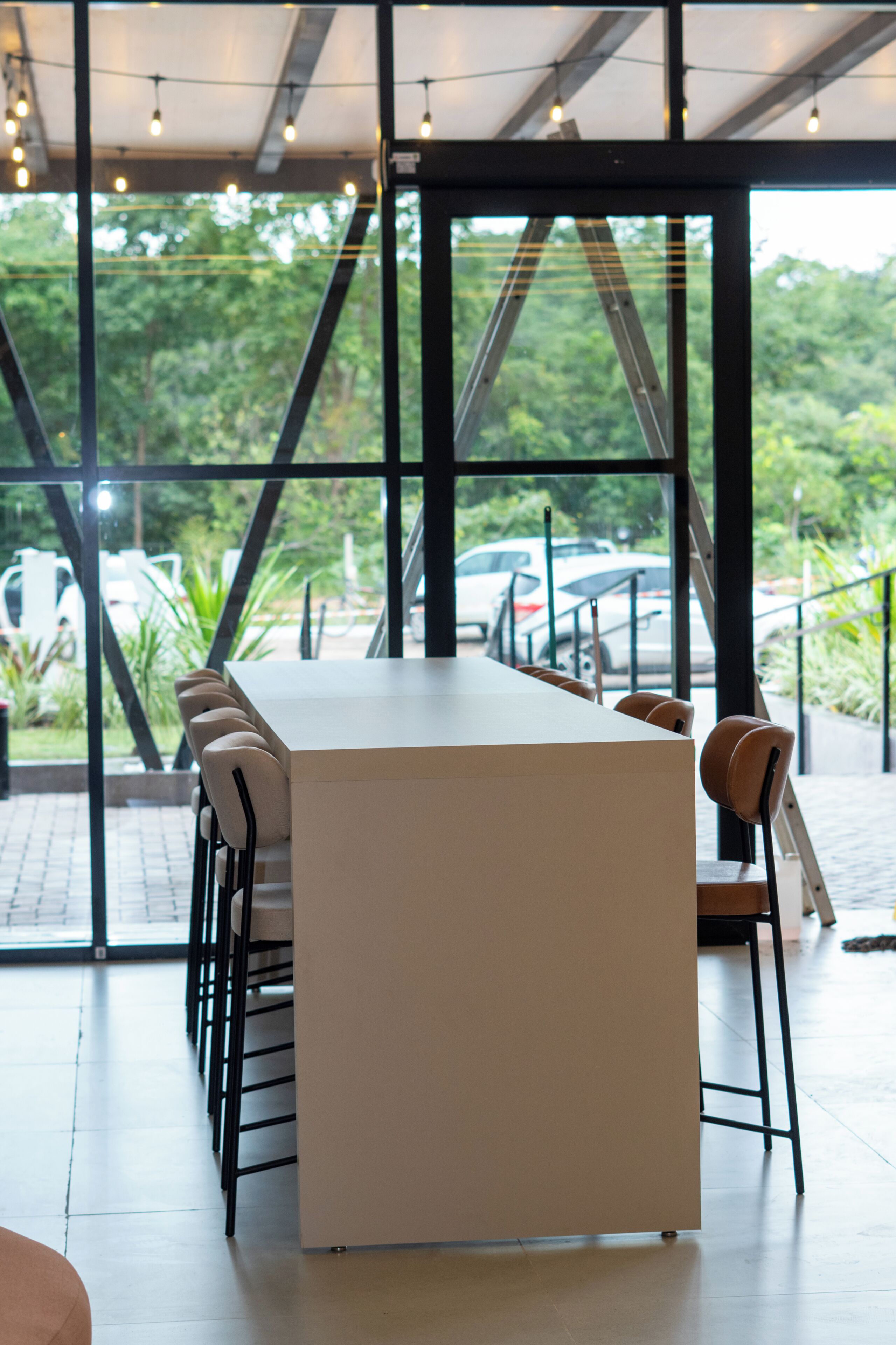 A modern bar area with a long counter and multiple chairs, large windows with a view of greenery and parked cars outside.