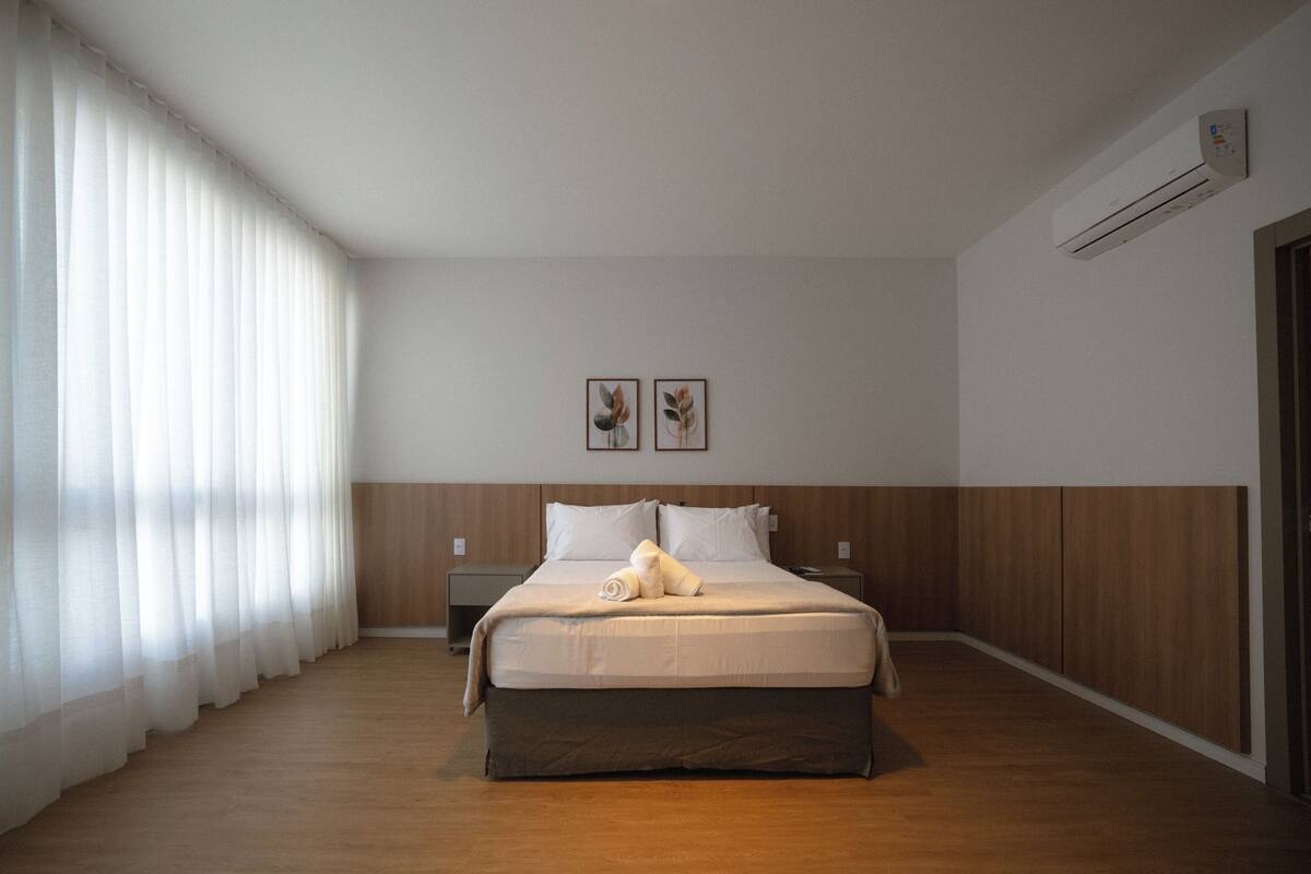 Une chambre d’hôtel avec un grand lit, un plancher en bois, des rideaux blancs et un appareil de climatisation fixé au mur.