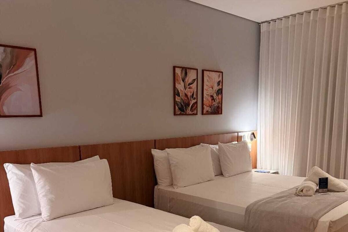 Une chambre d’hôtel avec deux lits, une tête de lit en bois et une grande fenêtre avec des rideaux blancs.