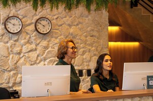 Dos recepcionistas en un mostrador con computadoras, una pared de piedra y relojes al fondo.