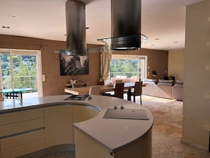 A modern kitchen with a curved island, a dining area with a glass table, and a view of the outdoors. - Splendid Villa with swimming pool (Roquefort-les-Pins)