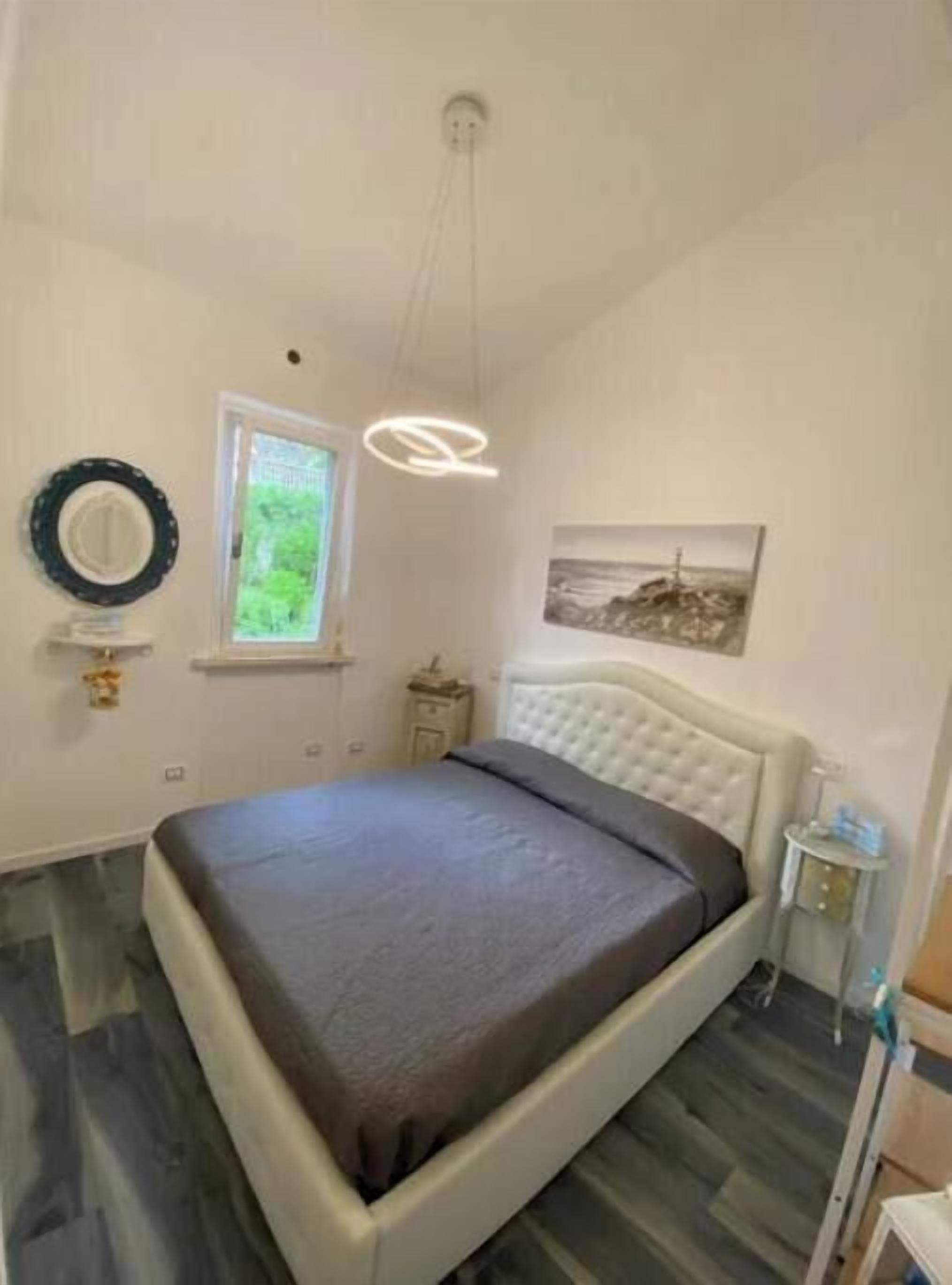 A bedroom with a bed, a window, a wall-mounted mirror, a picture, and a ceiling light fixture.