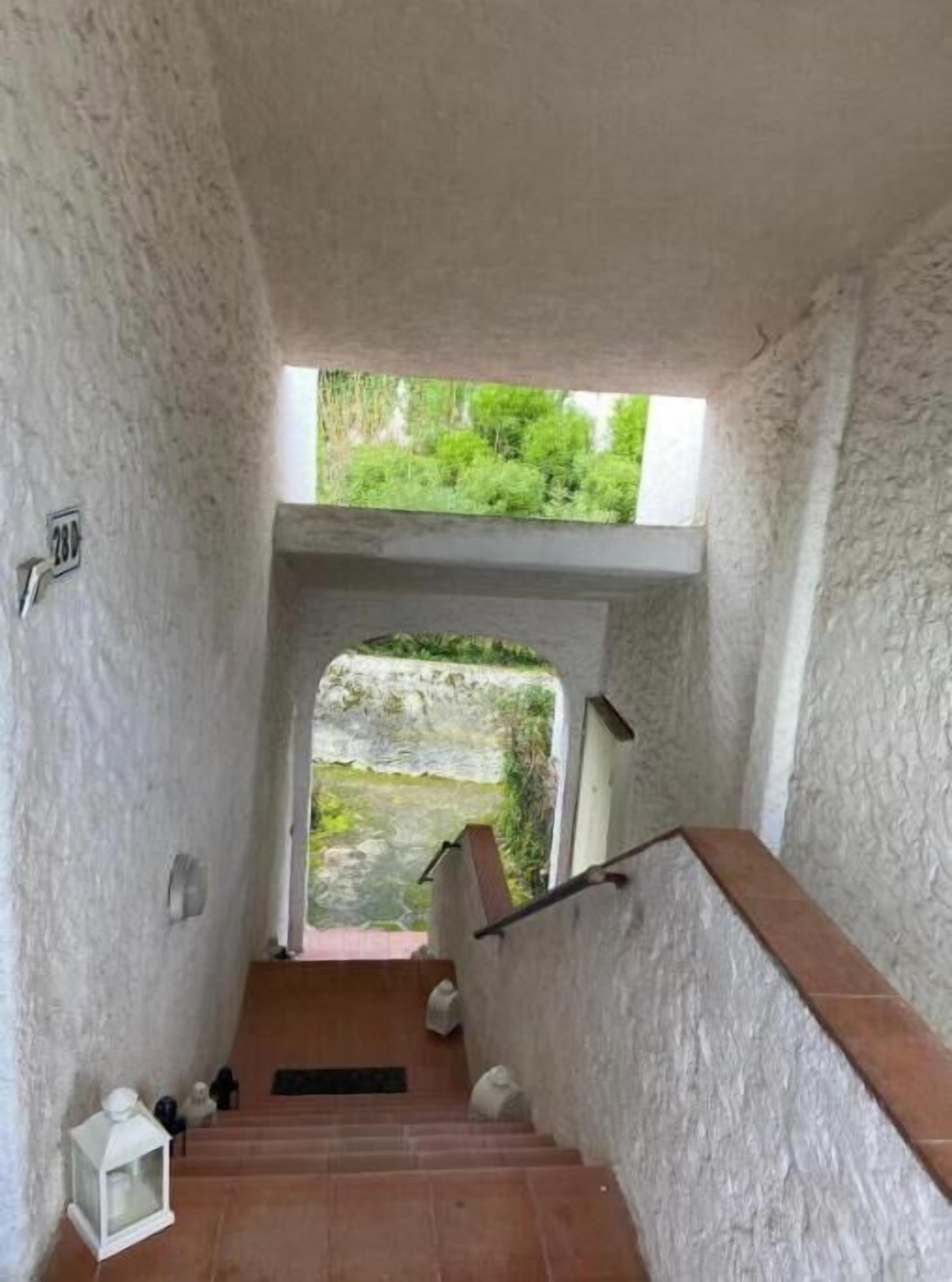 Un escalier avec un mur blanc, une petite lanterne et une vue sur la verdure à l’extérieur.