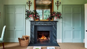 A room with a fireplace, two doors, a chair, a basket of firewood, and a table with books.