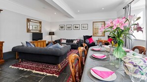 A modern living room with a sofa, ottoman, dining table set for four, and a large window with curtains. - Welsh Coach House - Five Bedroom Villa, Sleeps 9 (Tan-y-groes)