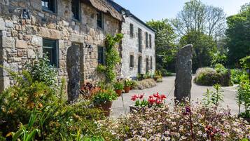 A stone building with a thatched roof, surrounded by a garden with various plants and flowers.