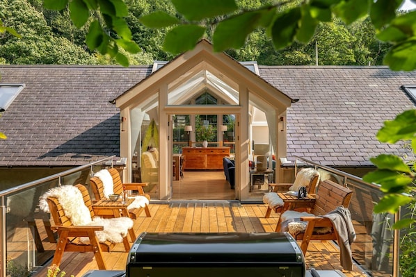 A wooden deck with outdoor furniture, a glass-enclosed dining area, and a view of a forested area.