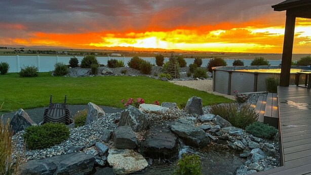 Un jardín ajardinado con terraza de madera, una parrilla y vistas a un atardecer sobre el agua.