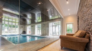A modern indoor pool with glass walls, a stone wall, and a beige sofa. - Pendragon Manor - Nine Bedroom Villa, Sleeps 18 (Cilybebyll Pontardawe)