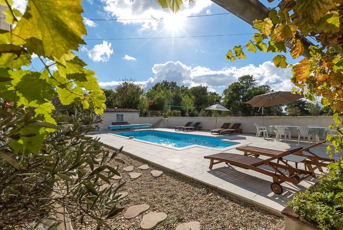 Piscina con sillas de descanso y una mesa, rodeada por un patio con vistas a árboles y un edificio al fondo.