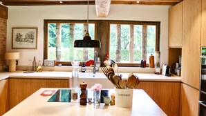 A modern kitchen with wooden cabinets, a large island, a hanging light, and a window with a view of trees. - La Faisane - Five Bedroom Villa, Sleeps 10 (Épaignes)