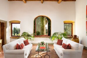 Ein geräumiges Wohnzimmer mit zwei weißen Sofas, einem Glastisch und großen Fenstern mit Rundbögen.