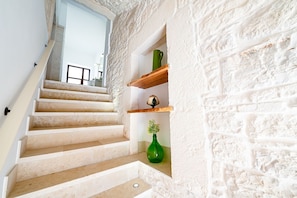 Eine Treppe mit beigen Stufen führt zu einem Raum, dessen Wand weiß und strukturiert ist und auf einem Regal eine grüne Vase steht.