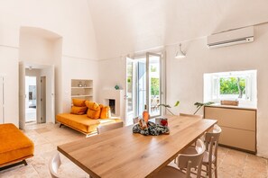 Ein modernes Wohnzimmer mit einem Holzesstisch, einer Couch mit orangenen Kissen, einer Küchenzeile und Blick ins Grüne.