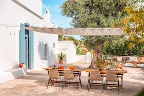 Eine Terrasse mit Tisch und Stühlen, ein weißes Gebäude mit blauen Fensterläden und ein Baum.