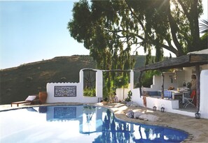Ein Pool mit weißer Wand und einem mosaikartigen Fliesenbelag, umgeben von Gartenmöbeln und einem Essbereich.