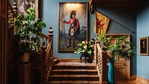 A grand staircase with ornate railings, a large framed portrait, and lush green plants.