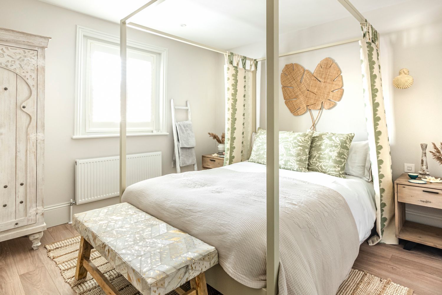A bedroom with a four-poster bed, a bench, a radiator, a window, and a bedside table with a lamp.