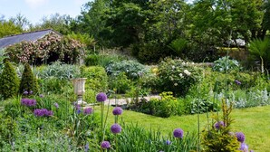 A garden with a variety of plants and flowers, a stone structure, and a building with a sloped roof.