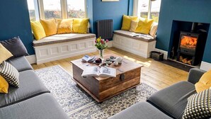 A living room with a fireplace, seating with yellow cushions, a coffee table with books and a vase, and a view through large windows. - Spinnaker House - Eight Bedroom Villa, Sleeps 16 (Beaumaris)