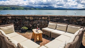 A patio with wicker furniture and a stone wall, overlooking a body of water with boats and mountains in the distance. - Spinnaker House - Eight Bedroom Villa, Sleeps 16 (Beaumaris)