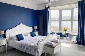 Un dormitorio con una cama grande, una mesita de noche, una silla, una ventana con vistas al mar y vista al mar a través de la ventana.