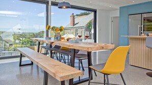 Un moderno comedor con una mesa de madera, sillas y un banco. Se aprecia una vista a través de amplios ventanales, una lámpara colgante y una zona de cocina con mostrador.