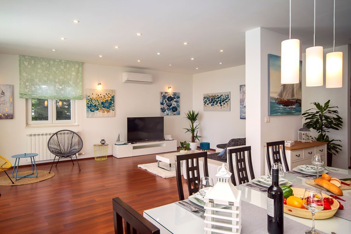 Una sala de estar moderna con televisor de pantalla plana, una mesa de comedor lista para dos personas y una cocina a la vista en el fondo.