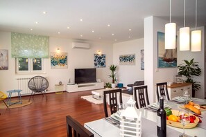 Ein modernes Wohnzimmer mit Flachbildfernseher, Esstisch für zwei Personen und einer im Hintergrund sichtbaren Küchenzeile.