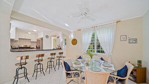 A dining area with a glass table, wicker chairs, and a view of the kitchen. - 3BR Townhouse in Holetown w/pool, Steps from Beach! (Holetown)