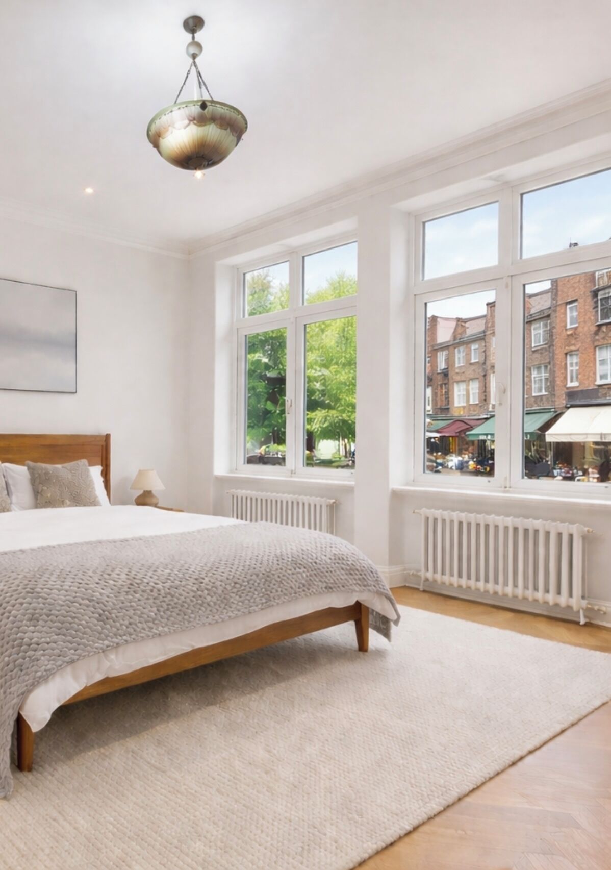 A bedroom with a large bed, a radiator, a window with a view of buildings, and a hanging light fixture.