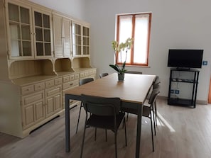 Ein Essbereich mit Holztisch und Stühlen, einem Blumentopf, einem Fernseher auf einem Schrank und einem Fenster mit Jalousien.