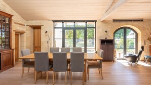 A dining area with a wooden table and chairs, large windows, and a stone wall.