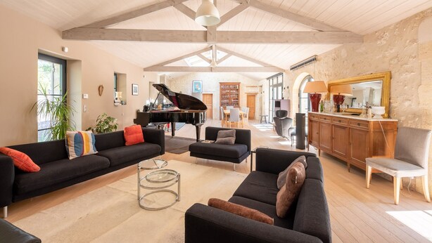A spacious living room with a grand piano, sofas, a dining area, and a kitchen with a large mirror.