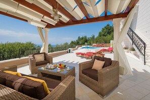 Ein überdachter Außenbereich mit Korbmöbeln, einem Pool und Meerblick.