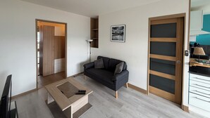 A modern living room with a sofa, a coffee table, a TV, and a door leading to a kitchen. - Urban cocoon with panoramic view - Balcony - Fully equipped (Saint-Étienne)