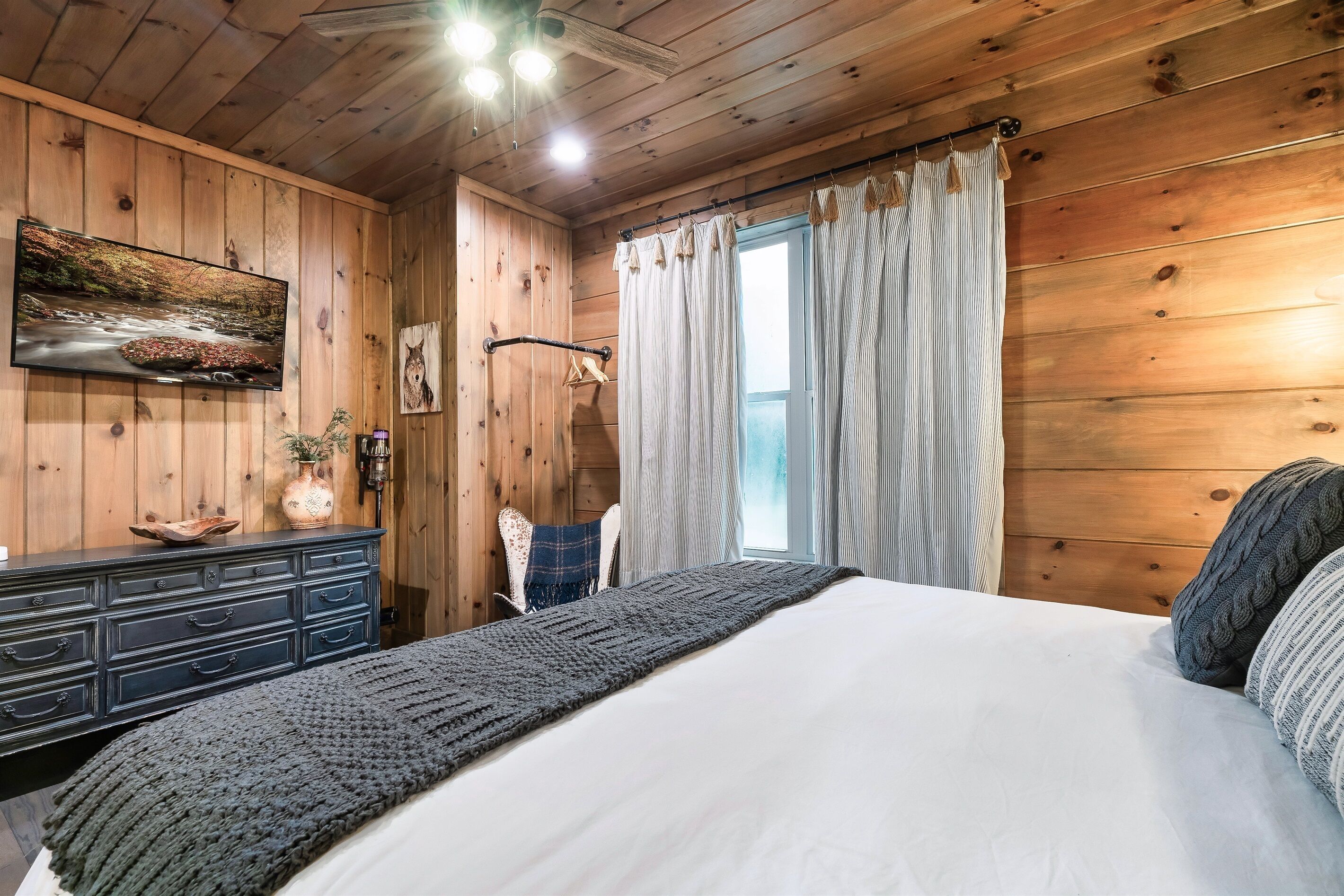 A wooden-paneled bedroom with a bed, a dresser, a painting, and a window with curtains.