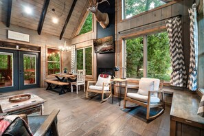 A rustic interior with wooden floors, a fireplace, and large windows overlooking greenery. - Luxe Craftsman | 3 King Suites | Fire Pit | Top 5% (Gatlinburg)