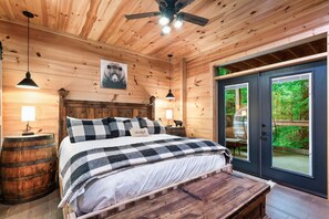 Un dormitorio de cabaña de madera con una cama, dos lámparas de noche, un ventilador de techo, un banco de madera y un ventanal con vistas a la vegetación.