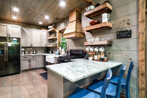 A kitchen with wooden cabinets, a stainless steel refrigerator, a marble countertop island, and blue chairs. - Luxe Craftsman | 3 King Suites | Fire Pit | Top 5% (Gatlinburg)