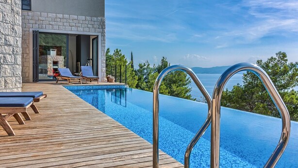 En modern infinitypool med ett trädäck, solstolar och en yttervägg av sten.