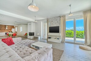 Ein modernes Wohnzimmer mit einer großen Eckcouch, einem Flachbildfernseher an einer Steinwand und einem Essbereich im Hintergrund.