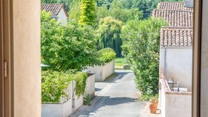 Blick durch ein Fenster auf eine enge Straße mit Häusern, Grünflächen und blauem Himmel.