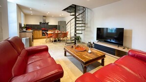 Uma sala de estar moderna com sofás de couro vermelho, uma mesa de centro de madeira, uma televisão e uma cozinha ao fundo.