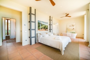 Quarto com cama, janela grande, pintura, ventoinha no teto e banheiro com banheira.