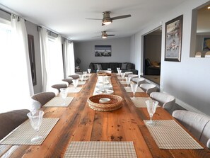 Una larga mesa de comedor de madera con múltiples sillas, un ventilador de techo y un cuadro en la pared.