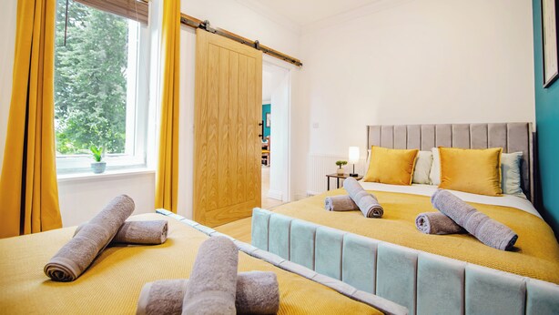 Habitación de hotel con dos camas, un ventanal con cortinas, una puerta de madera y vistas a zonas verdes.