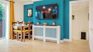 Una sala de estar con un televisor montado en una pared azul verdoso, una mesa de comedor de madera con sillas y una consola blanca.