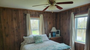 Un dormitorio con paredes de madera, una cama con ropa de cama floral, un ventilador de techo y una ventana con cortinas.