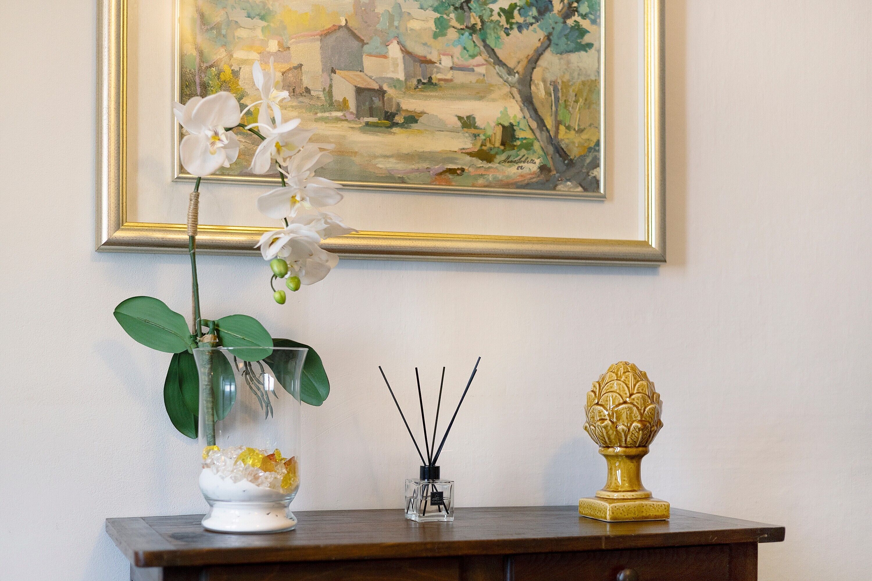 A vase with an orchid, a small glass container with reed sticks, and a decorative urn on a wooden table against a wall with a framed painting.